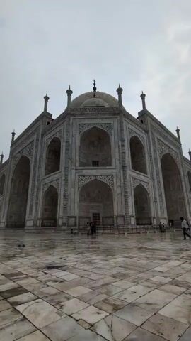 The video shows a low-angle, vertical shot of the Taj Mahal. The sky is overcast, and the marble facade of the monument is visible, with its intricate carvings and arches. People are walking on the tiled plaza in front of the Taj Mahal. In the distance, a minaret is visible, as well as a body of water. The ground is wet, suggesting it may have rained recently.