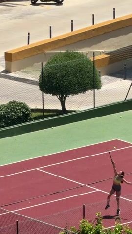 A long shot shows a woman playing tennis on an outdoor court. The court is painted red with green borders and white lines. The woman, wearing a dark grey and yellow athletic outfit with red hair, is hitting a tennis ball with a green and black tennis racket. There are multiple tennis balls on the court. A black chain link fence surrounds the court. The background shows a parking lot with cars and various trees and shrubbery. The scene is bathed in sunlight.