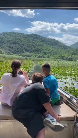 The video shows three children from behind, seated on the back of a boat, looking out at a calm river with water lilies and reeds. A lush green mountain covered in trees rises in the background under a blue sky with white clouds. The children appear to be on a leisurely boat ride.