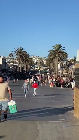 The video captures a bustling outdoor scene on a sunny day, likely a boardwalk or promenade, with a clear blue sky overhead. In the foreground, a person walks away from the camera carrying a teal and white cooler. The middle ground is populated by a crowd of people walking in both directions along a wide, paved walkway lined with palm trees. Shops and buildings with awnings are visible on either side of the walkway. A person on a bicycle is also present in the middle ground. The background features more buildings and palm trees, suggesting a coastal town or resort. The right side of the frame shows a portion of a light brown brick wall with two bronze plaques mounted on it. The plaques appear to be commemorative or informational, with text and a city seal visible on one. The overall impression is one of a lively, public space during daytime.