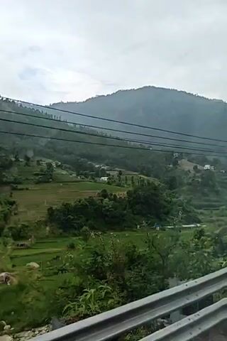 The video shows a scenic view of lush green hills and valleys. The terrain is terraced with patches of what appear to be rice paddies or agricultural fields. Trees and vegetation cover the slopes, and a cloudy sky looms overhead. Power lines are visible crisscrossing the landscape. The overall impression is of a rural, mountainous area.