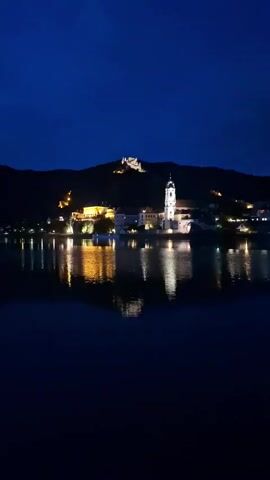 A still, vertical nighttime shot shows a small town nestled along a body of water, likely a river or lake. The town is illuminated by warm, artificial lights that reflect on the dark water. Dominating the skyline is a prominent white church steeple, its cross visible against the deep blue night sky. Behind the church, a hill rises, topped by the silhouette of ancient castle ruins. Buildings with illuminated windows line the waterfront, suggesting businesses or homes. The water itself is calm, creating a mirror image of the town's lights and architecture. The overall atmosphere is tranquil and picturesque. There are no people or discernible actions in the video.