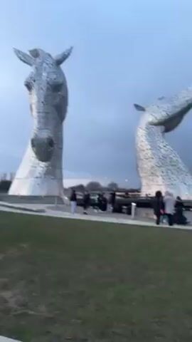 The video shows two large, silver, metallic horse head sculptures in a park-like setting, with people walking around and taking pictures. The sculptures are set against a cloudy sky. The people are diverse in age and dress and appear to be tourists or visitors. There is a water feature around the base of the sculptures. The grass is green and slightly unkempt. Street lamps are visible in the background. The overall lighting suggests it's either early morning or late afternoon.