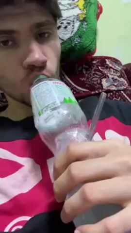 A fair-skinned young man with brown hair, wearing a red and black shirt, is seen from the chest up, holding a clear plastic bottle to his mouth. The bottle is partially filled with a clear liquid, and a clear straw is inserted into the side. He appears to be drinking from the bottle, making sucking noises. The background consists of a patterned cushion and a green decorative item with a cartoon image. The setting appears to be indoors, possibly on a sofa.
