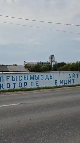 A wide shot of a road and a long white wall with blue lettering. The wall is covered in text, some of which is visible and some of which is obscured by trees. The text appears to be in Russian. Buildings are visible behind the wall, and there are trees lining the wall. The road is paved and has white lane markings. A silver car drives by on the road, and two people walk on the side of the road. The sky is overcast.