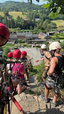 The video shows a group of people at the edge of a cliff, preparing for a zipline activity. In the foreground, several individuals are wearing helmets and harnesses. One person, on the right, is guiding the participants and appears to be an instructor. They are holding a rope and the other individuals are holding onto the zipline equipment. In the background, there is a village with buildings, some with red roofs, nestled in a green, hilly landscape. The sun is shining brightly, indicating a clear, sunny day, likely midday given the lighting. The overall atmosphere seems adventurous and exciting, with the participants appearing ready for the thrill of the zipline. Visible text on the white helmet of the instructor includes "OUTDOOR" and possibly a brand name or logo. The scene is set in Warisy, Belgium, suggesting this is a popular tourist or recreational spot for outdoor adventures.