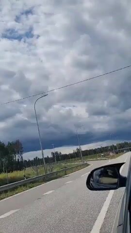 The video shows a view from the passenger seat of a car traveling on a highway. The road is paved and has white lane markings. On the left side of the road, there is a grassy area with some trees and a metal fence. There are also streetlights and traffic signs visible. In the distance, there are more trees and some construction activity. The sky is cloudy, with some dark clouds suggesting rain. The car's side mirror is visible in the foreground, reflecting the sky and the road. The video appears to be filmed while moving, as indicated by the blur of the passing scenery.