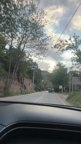 The video shows a view from inside a car driving on a road. The road is flanked by greenery and buildings. A few cars are visible in front of the car, and a security checkpoint with lit up green bars can be seen in the distance. The sky is cloudy and the sun is shining through the clouds. The car appears to be moving at a steady pace. Music is playing in the background.