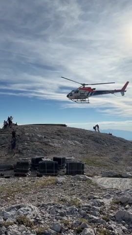 A helicopter flies over a rocky, mountainous terrain. Several people are visible on a ridge, some of whom appear to be walking. In the foreground, several large, dark rectangular objects are arranged on pallets. The sky is blue with scattered clouds, and the sun is visible.