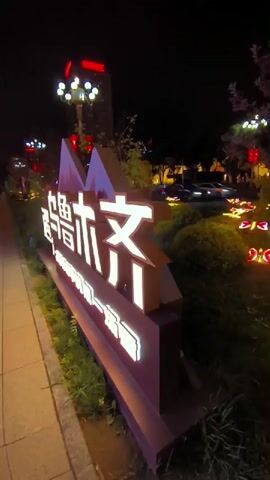 A nighttime outdoor scene shows a sign with illuminated Chinese characters, possibly marking an entrance or location. The sign is lit in white and sits in front of a planter bed filled with green bushes and grass. The bushes are decorated with small, colorful illuminated figures that resemble butterflies or lanterns, casting a warm glow on the foliage. A tree with purple-tinged leaves stands behind the planter, and more lights are visible in the background, suggesting a street or pathway with traffic. The overall impression is festive and decorative, likely set up for an event or as part of a city's nighttime illumination.