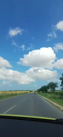 The video shows a view from inside a vehicle driving on a paved road through a rural landscape. The sky is bright blue with scattered white, puffy clouds. On either side of the road are fields, some appearing to be crops and others grassy. Trees line the right side of the road. In the distance, two motorcycles and a red truck are visible on the road. The vehicle appears to be moving at a moderate speed. The overall impression is a sunny day in the countryside.