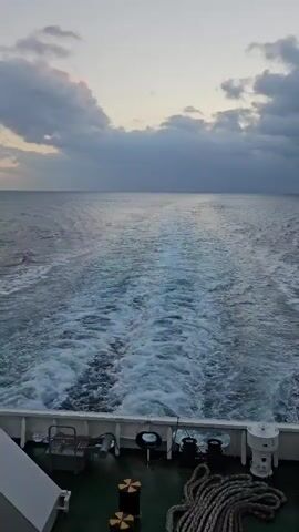 The video shows a view from the stern of a ship sailing through the ocean. The water behind the ship is churned up into a white wake. The sky is overcast with some patches of pinkish clouds visible. The deck of the ship is visible in the foreground, with various pieces of equipment, including winches and coiled ropes. The overall scene suggests a journey at sea, possibly during sunrise or sunset.