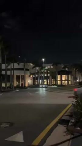 At night, a wide paved road leads towards a large, brightly-lit building, framed by numerous tall palm trees. Two cars are driving on the road, their headlights shining clearly. The building has a modern architectural style and features multiple windows that emit a warm, yellow glow. The sky above is dark. The scene is captured from a static position, with a sidewalk and a small patch of greenery visible in the foreground on the right side.