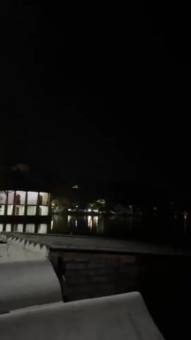 The video shows a nighttime view of a lake. A building with illuminated windows is visible on the left. Across the lake, lights from other buildings and structures are reflected in the water. The camera is positioned near a low wall or ledge, partially obscuring the view. The scene is dark overall.