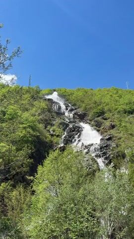 The video shows a waterfall cascading down a rocky hillside, surrounded by lush green trees and foliage. The sky is a clear, bright blue. Power lines are visible in the upper portion of the frame. The waterfall is multi-tiered, with water spraying into the air at the top.
