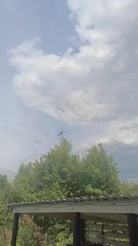 A helicopter is captured in flight against a backdrop of a cloudy sky. The helicopter appears to be a medium-sized, military or transport type, with visible rotors and landing gear. It is positioned in the upper portion of the frame, moving across the sky. Below the helicopter, the tops of lush green trees are visible, suggesting a rural or suburban setting with abundant vegetation. In the foreground, a portion of a wooden structure, possibly a roof or an overhang with visible beams and supporting posts, partially obscures the lower part of the scene. The lighting suggests it is daytime, with the sun filtering through the clouds, creating a mix of bright and shadowed areas in the sky. The presence of clouds indicates a potentially variable weather condition. The location is identified as Zubovo, Russia. No people are visible in the frame. The overall mood is one of observation, with the focus on the airborne object and the natural surroundings. There is no visible text within the media file.