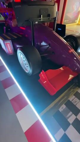 A purple and gold racing simulator is displayed with a screen mounted on top. A person sits in the driver's seat. Several people stand around the simulator, some looking at the screen and others interacting with each other. The area appears to be an indoor exhibition or event space, with checkered flooring and red and white striped barriers. Neon blue lights illuminate the simulator.