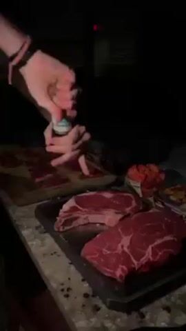 A person stands at a kitchen counter in a dimly lit room, preparing food. Their hands are visible as they grind a black pepper mill over two large, raw steaks on a dark grey baking sheet. To the left, there's a wooden cutting board with several thin slices of raw meat. Other items on the counter include a container of chopped tomatoes and a small cardboard package, possibly containing snacks. The person wears a wristband with alternating black and pink bands. The background is dark, but there's a window with closed blinds and a small red indicator light visible.