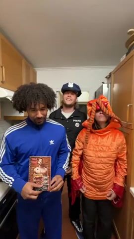 Here is a detailed description of the video:

Three people stand in a kitchen. The kitchen has wooden cabinets and white walls with a white ceiling. The person on the left is wearing a blue tracksuit with white stripes. They hold up two items in front of them, one at a time. The first is a magazine with a man on the cover. The second is a white card with the words "Et voici Georges charlie de livrer son gateau." A photo of a man holding a cake is in the middle. The card is covered in handwritten notes. The person in the center is wearing a police costume. The person on the right is wearing an orange costume with large, red bug eyes and long, orange tentacles.