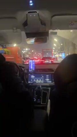 The video is shot from the inside of a car, likely the back seat, while it's stuck in traffic at night. The weather appears to be snowy or rainy. We see the dashboard of the car, featuring a digital display showing a radio station. There's also a smartphone mounted, displaying a map. Through the windshield, we see numerous other cars and a large truck ahead, their brake lights illuminating the scene. A person is visible walking between cars. The overall setting seems to be an urban environment.
