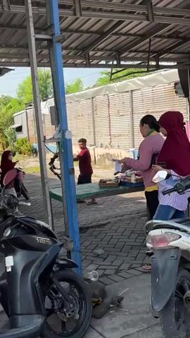 Two women are seen at an outdoor market stall. One woman, wearing a burgundy hijab and a striped shirt, is facing away from the camera, looking at goods on display. The second woman, wearing a pink top, is standing next to her, also looking at the merchandise. In the foreground, a white scooter is parked. In the background, a second stall is visible with various food items displayed on tables. Another woman wearing a black hijab and a white top is standing at this second stall, interacting with a young boy who is also looking at the goods. The market appears to be open-air with a covered roof structure.