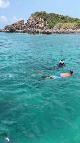 The video shows three people snorkeling in clear turquoise water near a rocky, green island. One snorkeler is wearing a black wetsuit and mask. Another is wearing blue swim trunks, an orange shirt, a mask, and a snorkel. A third snorkeler is wearing a black mask and a black top, with what appears to be a camera or a phone attached to their arm. All three are swimming and appear to be looking at something underwater. The sky is clear and blue.