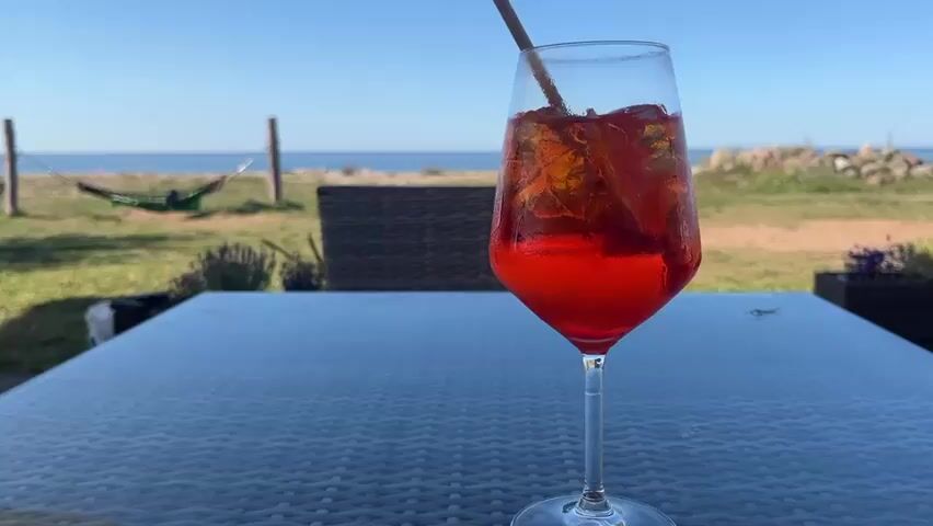 A close-up shot focuses on a glass of red liquid with ice and a straw, placed on a dark, textured table. In the background, a body of water, likely the sea, is visible under a clear blue sky. A green hammock is slung between two poles on a grassy area to the left. A chair is partially visible on the right. The overall setting appears to be an outdoor space, possibly a cafe or restaurant, with a view of the coast.
