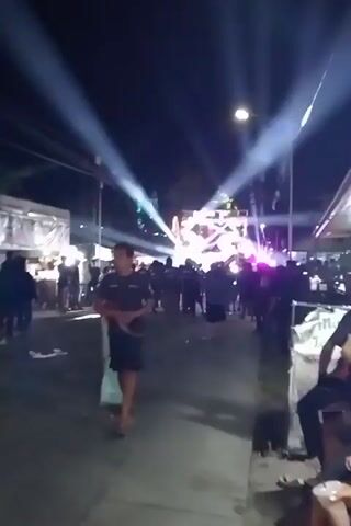 The video shows a nighttime outdoor event. A crowd of people is gathered in front of a stage or setup with bright lights. The lights are beaming upwards and creating a dramatic effect in the dark sky. There are tents or stalls set up on either side of the area where the crowd is standing, suggesting a market or festival atmosphere. In the foreground, a few individuals are visible, some walking and others seated. The overall impression is one of a lively public gathering at night, possibly a concert or celebration, characterized by bright lighting and a significant presence of people.