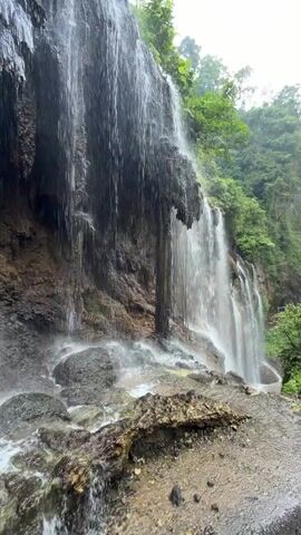 A waterfall cascades down a moss-covered cliff face surrounded by lush green foliage. The water crashes onto a rocky riverbed, creating a misty spray. The shot is taken from a low angle, looking up at the waterfall.
