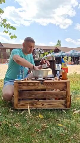 A man in a green t-shirt and blue shorts kneels on the grass outdoors. He is in front of a wooden crate that serves as a table. On the crate, there is a silver pot, a small white cup, a bottle of water, a coffee grinder, and two small flags - one white and red, and another blue, yellow, and red. The man is holding a coffee grinder with a small handle and appears to be grinding coffee beans. Behind him, there is a building with a brown roof and blue awnings, and some trees. The sky is blue with white clouds.
