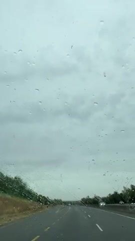 The video shows a view through a windshield covered in raindrops, looking out at a highway on a cloudy day. The road appears to be empty of other vehicles in the immediate foreground, but there are some vehicles visible in the distance. The sky is overcast and gray. On the left side of the road, there is a grassy embankment with some trees. On the right side, there is more vegetation. The road has yellow lines separating the lanes. The camera perspective suggests the video was taken from a moving vehicle.