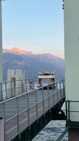 A ferry boat with passengers and vehicles is approaching a dock. The ferry is white with red accents and has a cabin on top. It is on a body of water with mountains in the background. The mountains are lit by the setting sun, casting a warm glow on their peaks. The dock is made of metal and has a ramp leading to it. The water is choppy and reflects the sky. The scene appears to be at sunset.