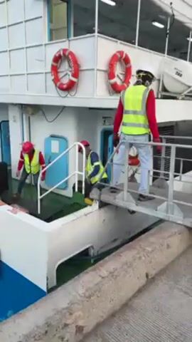 The video shows three people wearing safety vests and hard hats boarding a white boat. One person stands on a metal gangway leading onto the boat, while the other two are carrying luggage and walking up the steps. The boat has life rings attached to its side.  The boat is docked next to a concrete structure.