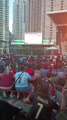 A large outdoor screen displays a soccer match, with fans gathered in front of it. The screen is positioned above a crowd of people sitting in chairs and at picnic tables, all facing the screen. The fans are dressed in casual clothing, with some wearing jerseys. Buildings rise in the background, suggesting an urban setting. The lighting indicates it is either dusk or early evening. The video captures a moment of shared experience among the fans as they watch the game.