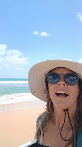 Here's a description of the video:
A woman is on a beach, taking a selfie video. She is smiling and holding a black boogie board under her arm. She is wearing a light-colored hat, sunglasses, a blue and white floral bikini top, and a green life vest. The beach has golden sand and turquoise water with waves crashing in the background. The sky is blue with scattered clouds. The woman appears happy and relaxed.