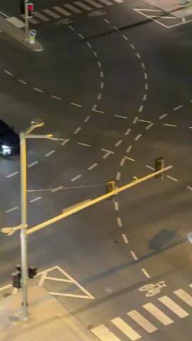 A black car turns at a street intersection at night. The intersection has traffic lights, pedestrian crossings, and lane markings. Another car is seen turning across the intersection. Another black car is driving down the road at speed, and a white car is seen driving past. The black car continues to drive down the road. Palm trees are visible on the side of the road.