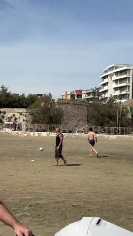 Two people are playing soccer on a sandy beach under a blue sky with patchy clouds. One is an adult male wearing a black tank top and dark shorts, with visible tattoos on his arms and calves, and he is walking on the sand. The other is a younger male, shirtless and wearing dark blue shorts, who is running and kicking a soccer ball. The beach is surrounded by a low stone wall and trees, and in the background, there are buildings and a lamppost. A baby carriage is visible in the foreground.