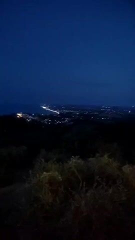 The video shows a dark, panoramic view of a coastal town at night from a hilltop. The sky is a deep blue, transitioning to a lighter blue near the horizon where the ocean meets the land. The town lights sparkle along the coastline and spread inland, creating a gentle glow against the darkness. The foreground is filled with shadowy bushes and dry vegetation, adding depth to the scene. The camera appears to be moving slightly, causing a subtle blur.