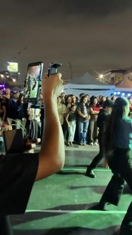 A group of people are dancing on a stage at night. The stage is lit with purple and green lights. A crowd of people are watching the performance from behind a railing. One person in the foreground is holding up a phone to record the performance. There are tents and streetlights in the background.