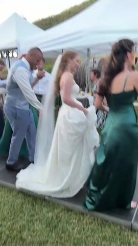 A group of approximately ten adults, including a bride and groom, are engaged in a line dance, holding hands on a raised black outdoor dance floor. The bride, wearing a white gown and veil, leads the line to the left of the frame, followed by the groom in a light blue vest and white shirt, and several guests in various attire, including green dresses. In the background, multiple white tents are erected on a grassy area, with a natural incline visible further back. String lights are strung under the tents.