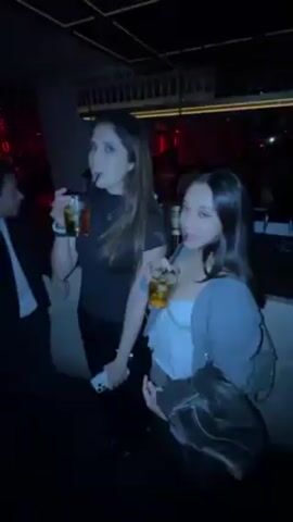 Two women are standing in a dimly lit bar, each holding a drink. The woman on the left is older, with long dark hair and a black top, while the woman on the right is younger, with a white top, gray jacket, and shoulder bag. They both take sips from their drinks, moving slightly to the music. The bar has bottles displayed on shelves in the background. There are other people in the bar as well. The lighting is dark, with red accents.