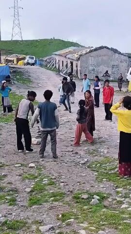 In a rural, mountainous setting with scattered dilapidated buildings and a visible power line tower, a group of children and adults are gathered. The ground is uneven and covered with rocks and sparse vegetation. Several children appear to be engaged in some form of play or activity. One girl in the foreground, wearing a dark t-shirt and orange pants, looks towards the camera. To her right, another girl in a yellow long-sleeved shirt and a long patterned skirt stands facing away from the camera. In the background, a cluster of people, including children and adults, are interacting. Some of the buildings have corrugated metal roofs and appear unfinished or in disrepair. Two white vehicles are visible in the background, one of which is a van. The overall impression is a scene of community life in a humble, possibly remote, location.