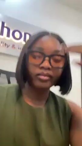 A woman with a dark complexion and short, straight black hair, wearing a muted green sleeveless top and large, square-framed eyeglasses with tinted lenses, is seated and looking directly at the camera. Her right hand is placed on her forehead, suggesting she is thinking or perhaps experiencing mild stress. Behind her, slightly out of focus, is a sign with the word "sona" visible, followed by "ity & inter". The setting appears to be an office environment.