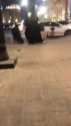 The video shows a bricked area outdoors, possibly a street or plaza, at night.  Several people in dark clothing are walking in the distance.  Two white cars are parked nearby.  The camera appears to be low to the ground and moves slightly.  The scene is lit with a warm light source.