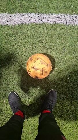 The video shows a person kicking a soccer ball on a soccer field. The person is wearing black sneakers and black pants with a red stripe. The soccer ball is old and worn, with visible scuff marks. The field is green with white lines, and the background shows a soccer goal and bright lights illuminating the field.