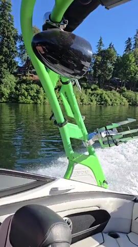 A first-person perspective video shows a boat moving across a body of water. The camera is positioned looking towards the stern of the boat, focusing on a green wakeboard tower with a black speaker attached. The boat is creating a substantial wake as it travels. Lush green trees and vegetation line the shore in the background, with houses visible amongst the trees. The sky is a clear, bright blue. The video captures the motion of the boat and the surrounding environment.