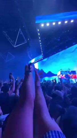 The video shows a lively indoor concert or music festival from the perspective of someone in the audience. A large crowd of people, mostly silhouetted against the bright stage lights, are enthusiastically cheering, dancing, and recording the performance with their mobile phones, held high in the air.

The stage in the background is brightly lit with vibrant blue and cyan lights, featuring a large screen displaying abstract visuals, possibly depicting a landscape with mountains. Performers, likely a DJ or band, are visible on the stage, which is also adorned with various lighting fixtures and structures. Triangular light fixtures or decorative elements hang from the ceiling on the left side of the frame, adding to the dynamic visual experience. The overall atmosphere is energetic and exciting, with the crowd fully engaged in the music. The location is Palm Desert, United States.