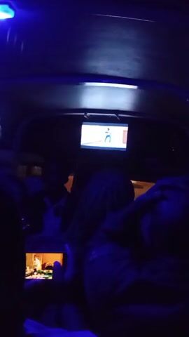 The video appears to have been taken inside of a vehicle, illuminated by a blue light. Several people are visible as silhouettes. There's a small television mounted inside the vehicle playing a video. Someone is also holding a smartphone, recording another video, likely displayed on the phone's screen. The interior of the vehicle seems crowded.
