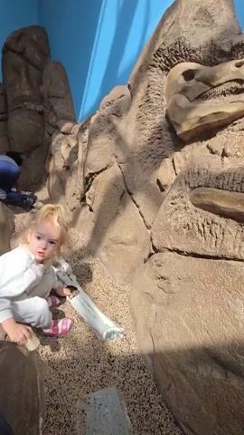 A young blond girl is crouching in a play area with simulated rock formations and a faux dinosaur skull. She's wearing a white long-sleeved top, light-colored pants, and pink sandals. She is playing with a short, silver, flexible tube. The ground is covered in small, multi-colored flecks of what appears to be synthetic material. Behind the girl are large rock-like structures that surround her. A dinosaur skull is embedded within the rock formation to her right. A glimpse of another person’s legs in jeans is visible behind her on the left. The background wall is painted a bright blue.