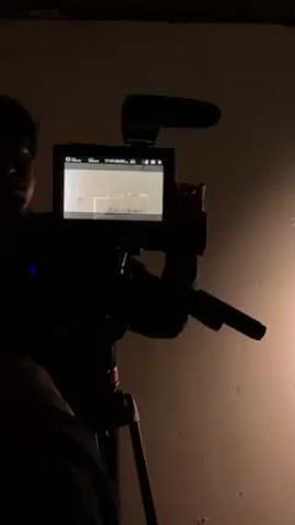 The video shows a person filming with a professional-looking camera on a tripod in a dimly lit room. The camera has a microphone attached and a screen showing the image being recorded. The person's face is partially visible, showing that they are wearing glasses. The wall behind them is textured and uneven, with a dark area at the top and a brighter area to the right.